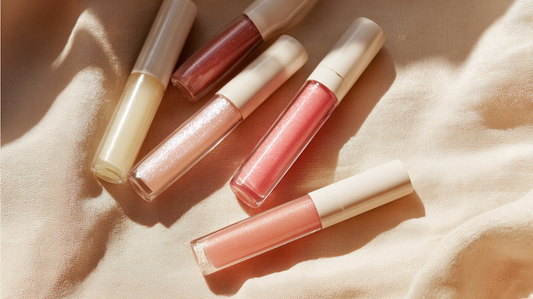 Essory Lip Gloss & Balms Collection featuring hydrating gloss tubes in nude, pink, and coral shades on a beige fabric background, moisturizing and glossy lip care for Fall 2025 makeup trends.